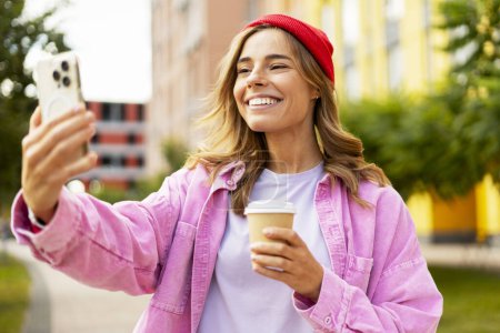 Téléchargez les photos : Femme souriante joyeuse portant un chapeau rouge tenant une tasse de café et prenant un selfie smartphone dans la rue. Influenceur utilisant la vidéo d'enregistrement d'application mobile, communication en ligne dans le parc - en image libre de droit
