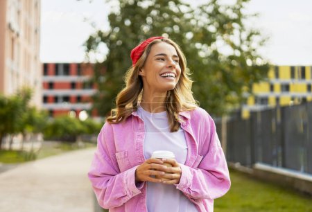 Téléchargez les photos : Belle femme positive aux cheveux ondulés portant un chapeau rouge élégant, des vêtements décontractés tenant une tasse de café regardant loin, voyageant dans la rue urbaine. Concept de pause café - en image libre de droit