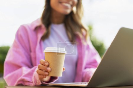 Téléchargez les photos : Gros plan d'une étudiante tenant une tasse avec du café, assise dans la rue, utilisant un ordinateur portable, un site Web, commandant. Concept d'achat en ligne - en image libre de droit