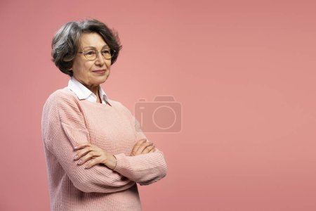 Foto de Retrato sonriente mujer mayor, abuela feliz vistiendo suéter, gafas con los brazos cruzados mirando hacia otro lado aislado sobre fondo rosa, espacio de copia - Imagen libre de derechos