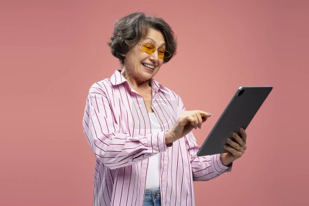 Téléchargez les photos : Portrait excité femme âgée, grand-mère heureuse tenant communication tablette numérique, achats en ligne, choisir quelque chose sur le site isolé sur fond rose. Concept technologique - en image libre de droit