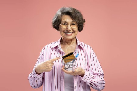 Foto de Mujer mayor de pelo gris sonriente, abuela feliz sosteniendo carro de la compra y tarjeta de crédito de oro aislado sobre fondo rosa. Compras, tienda, concepto de supermercado - Imagen libre de derechos