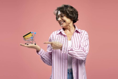 Photo for Smiling stylish senior woman, happy grandmother holding shopping trolley and pointing on golden credit card isolated on pink background. Shopping, store, delivery supermarket concept - Royalty Free Image