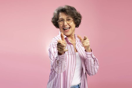 Foto de Mujer mayor emocionada, feliz abuela divertida con gafas y elegante camisa rosa bailando, divirtiéndose, señalando los dedos en la cámara aislada en el fondo - Imagen libre de derechos