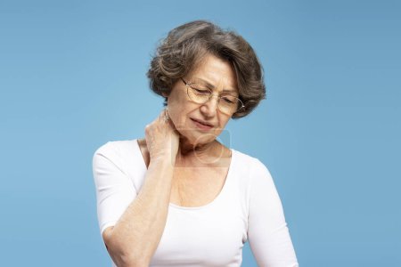Téléchargez les photos : Triste femme âgée ayant mal au cou se sentant mal isolé sur fond bleu. Soins de santé, concept de traitement - en image libre de droit