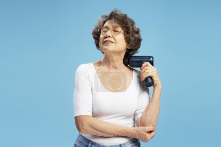 Téléchargez les photos : Heureuse femme âgée, grand-mère moderne à l'aide d'un pistolet de massage faisant auto massage du cou isolé sur fond bleu - en image libre de droit