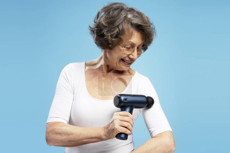 Photo for Smiling attractive senior woman, modern grandmother using massage gun for biceps, doing self massage isolated on blue background - Royalty Free Image