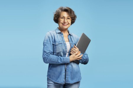 Foto de Hermosa atractiva mujer de negocios senior, feliz abuela moderna sosteniendo ordenador portátil mirando a la cámara aislada en el fondo azul. Negocios exitosos - Imagen libre de derechos