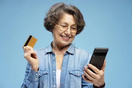 Téléchargez les photos : Femme âgée aux cheveux gris souriant, retraité heureux tenant carte de crédit et téléphone mobile faisant des achats en ligne isolé sur fond bleu. Services bancaires mobiles - en image libre de droit