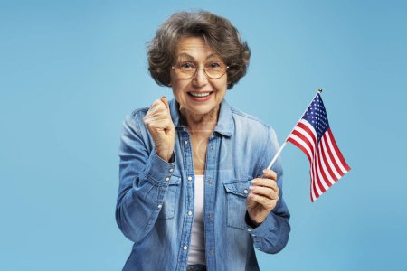 Photo for Smiling confident senior woman, happy modern grandmother holding American flag celebration Independence Day isolated on blue background. United States election concept - Royalty Free Image