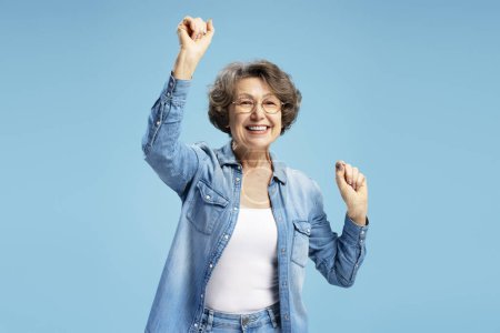 Téléchargez les photos : Attrayant souriant senior, heureuse grand-mère moderne tenant la main vers le haut célébration succès isolé sur fond bleu. Bonne nouvelle, concept de mode de vie positif - en image libre de droit