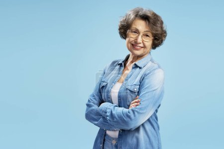 Foto de Mujer mayor sonriente con confianza en el retrato, abuela moderna feliz con ropa casual sosteniendo brazos cruzados aislados sobre fondo azul, espacio para copiar - Imagen libre de derechos
