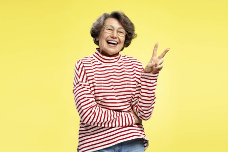 Téléchargez les photos : Belle femme âgée souriante portant un élégant pull à rayures rouges montrant le signe de victoire isolé sur fond jaune coloré. Style de vie positif - en image libre de droit
