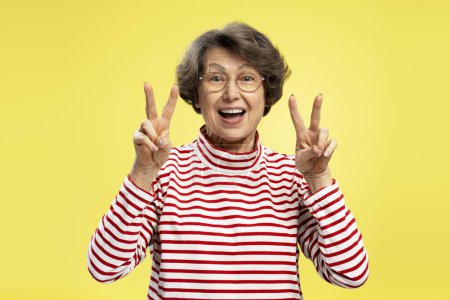 Photo for Funny smiling senior woman wearing stylish red striped sweater showing victory sign isolated on yellow colorful background. Positive emotions - Royalty Free Image