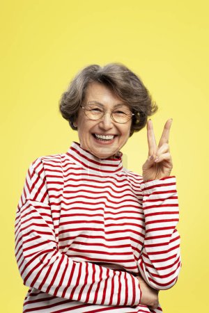 Photo for Smiling, mature woman wearing eyeglasses stylish sweater gesturing, showing with fingers peace sign isolated on yellow background. Concept of victory - Royalty Free Image