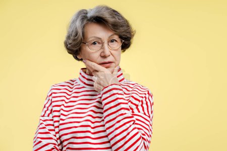 Téléchargez les photos : Pensionné sérieux, femme de 70 ans portant un pull élégant et des lunettes, regardant la caméra, choisissant quelque chose, debout isolé sur fond jaune. Concept de shopping, publicité - en image libre de droit