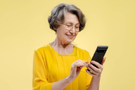 Téléchargez les photos : Portrait d'une séduisante femme aux cheveux gris portant des lunettes et des vêtements décontractés tenant un téléphone portable vérifiant le courrier bavardant debout isolé sur fond jaune. Concept de technologie en ligne - en image libre de droit