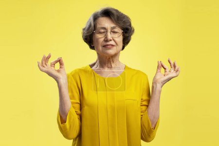 Téléchargez les photos : Femme âgée aux cheveux gris portant des lunettes avec les yeux fermés gesticulant, méditant debout isolé sur fond jaune. Concept d'harmonie, équilibre - en image libre de droit