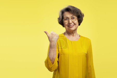 Foto de Retrato de una anciana positiva con anteojos apuntando con los dedos al espacio de copia mirando hacia otro lado aislada sobre fondo amarillo. Concepto de publicidad - Imagen libre de derechos