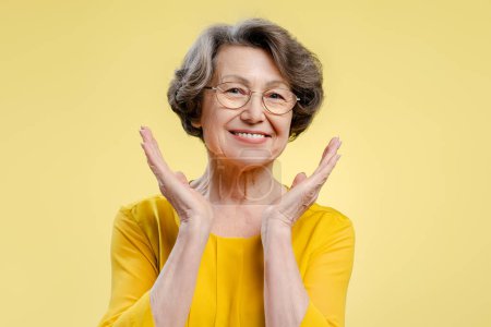 Foto de Retrato de una sonriente y hermosa mujer de los 70 con anteojos y una blusa amarilla casual, mirando a la cámara de pie aislada sobre un fondo amarillo. Concepto retirado - Imagen libre de derechos