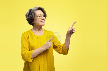 Foto de Retrato de una mujer mayor sonriente con anteojos apuntando con los dedos al espacio de copia mirando hacia otro lado aislada sobre fondo amarillo. Concepto de publicidad - Imagen libre de derechos