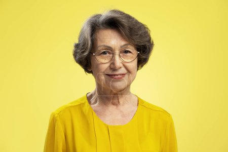 Téléchargez les photos : Jolie femme positive des années 70 portant des lunettes et un chemisier jaune décontracté, regardant la caméra debout isolée sur fond jaune. Concept de retraite - en image libre de droit