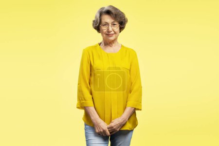 Foto de Mujer madura hermosa y sonriente con anteojos y blusa amarilla casual, mirando a la cámara de pie aislada sobre fondo amarillo. Concepto retirado - Imagen libre de derechos