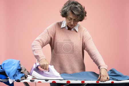 Téléchargez les photos : Portrait d'une femme âgée de 70 ans portant des lunettes tenant du fer à repasser sur une planche à repasser isolée sur fond rose. Concept de ménage - en image libre de droit