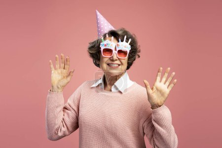 Foto de Sonriente hermosa mujer de 70 años de edad, vistiendo divertidas gafas con las manos en alto aislado sobre fondo rosa. Concepto de cumpleaños de celebración - Imagen libre de derechos