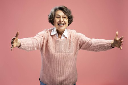 Téléchargez les photos : Portrait de belle grand-mère souriante, femme mûre portant des lunettes à bras ouverts, étreinte isolée sur fond rose. Concept d'amour - en image libre de droit