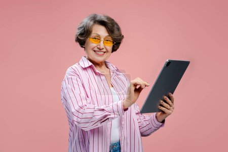 Téléchargez les photos : Happy senior femme aux cheveux gris hipster portant des lunettes jaunes élégantes et des vêtements décontractés tenant tablette numérique vérifier mail bavardage debout isolé fond rose. Concept de technologie en ligne - en image libre de droit