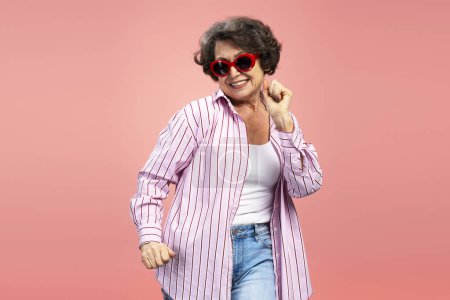 Foto de Retrato de mujer atractiva madura con gafas de sol bailando y mirando a la cámara aislada sobre fondo rosa. Concepto de publicidad - Imagen libre de derechos