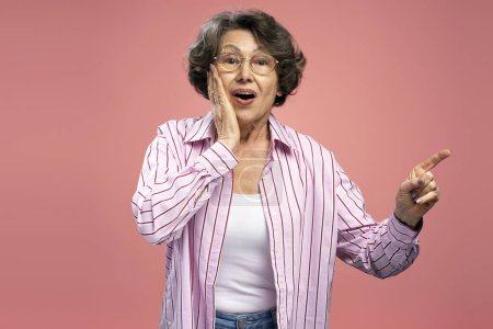 Foto de Emocionado hermosa mujer de 70 años madura con gafas, señalando el dedo en el espacio de copia de pie aislado en el fondo rosa. Concepto de compras - Imagen libre de derechos
