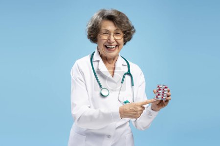 Téléchargez les photos : Portrait de médecin femme aux cheveux gris mûrs portant un manteau blanc tenant une plaquette thermoformée avec des pilules isolées sur fond bleu. Concept de traitement - en image libre de droit