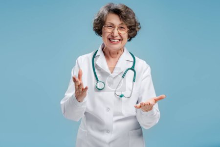 Téléchargez les photos : Portrait de femme âgée séduisante positive, médecin portant un manteau blanc, stéthoscope, regardant la caméra isolée sur fond bleu. Concept de soins de santé - en image libre de droit