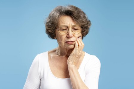 Téléchargez les photos : Portrait d'une femme âgée de 70 ans bouleversée portant des lunettes de vue touchant le mal de dents du visage isolé sur fond bleu. Concept de traitement, soins dentaires - en image libre de droit