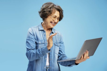 Téléchargez les photos : Portrait de femme aux cheveux gris souriant portant des lunettes à l'aide d'un ordinateur portable, ayant un appel vidéo, debout isolé sur fond bleu. Concept technologique - en image libre de droit