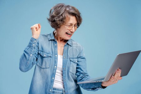 Téléchargez les photos : Portrait d'une femme aux cheveux gris de 70 ans souriante portant des lunettes à l'aide d'un ordinateur portable, gagnante, isolée sur fond bleu. Concept de paris sportifs, victoire - en image libre de droit