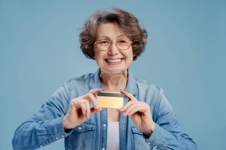 Téléchargez les photos : Portrait d'une séduisante femme âgée aux cheveux gris portant une carte de crédit, regardant la caméra isolée sur fond bleu. Concept de banque, publicité - en image libre de droit