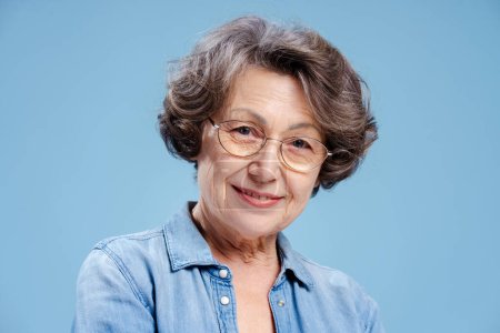 Téléchargez les photos : Portrait de femme âgée souriante de 70 ans portant des lunettes et une chemise en denim élégant isolé sur fond bleu, gros plan. Concept de publicité - en image libre de droit