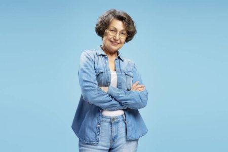 Téléchargez les photos : Portrait de femme âgée mature confiante portant des lunettes et une chemise en denim élégant, bras croisé isolé sur fond bleu. Concept de publicité - en image libre de droit