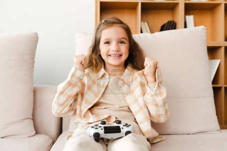 Téléchargez les photos : Portrait de belle petite fille gamer assis sur un canapé confortable jouant au jeu vidéo gagnant gestuel regardant la caméra s'amuser. Concept de jeu en ligne - en image libre de droit