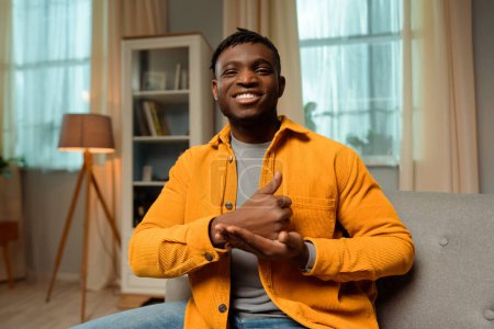 Téléchargez les photos : Portrait de souriant homme afro-américain heureux parlant avec la langue des signes, montrant des symboles de doigt, portant des vêtements décontractés, regardant la caméra tout en étant assis à la maison. Concept de communication - en image libre de droit