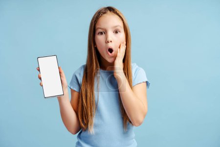 Téléchargez les photos : Portrait d'une petite fille surprise portant des vêtements décontractés tenant un téléphone portable avec écran vierge isolé sur fond bleu. Concept technologique - en image libre de droit
