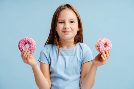 Foto de Retrato de una niña sonriente sosteniendo rosquillas, mirando a la cámara aislada sobre fondo azul. Concepto de comida, postre - Imagen libre de derechos