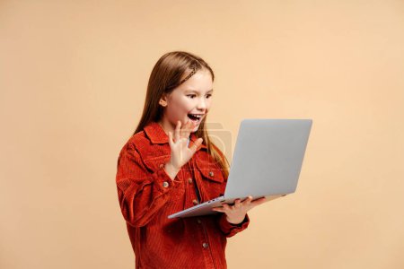 Téléchargez les photos : Portrait d'une belle fille souriante portant des vêtements décontractés, tenant un ordinateur portable, ayant un appel vidéo, agitant la main isolée sur fond beige. Concept technologique - en image libre de droit