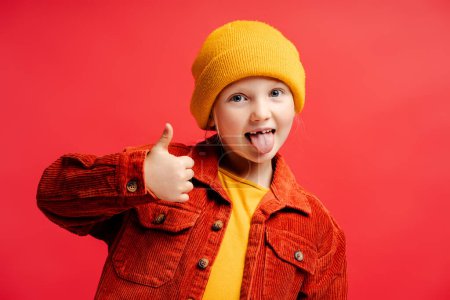 Foto de Retrato de niña linda con sombrero amarillo y ropa casual, mostrando el dedo grande, sonriendo de pie aislado sobre fondo rojo. Concepto de infancia - Imagen libre de derechos