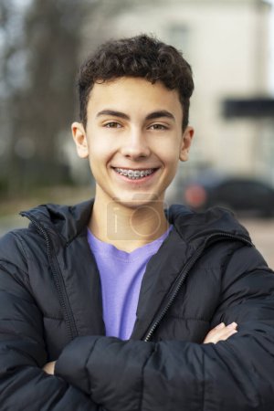 Foto de Retrato de niño guapo sonriente, adolescente con frenos con los brazos cruzados mirando a la cámara, usando chaqueta casual. Concepto de publicidad, cuidado dental - Imagen libre de derechos