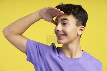 Photo for Portrait of attractive smiling boy with braces wearing stylish purple t shirt holding his hand to his forehead looking away, choosing something isolated on yellow background. Shopping concept - Royalty Free Image