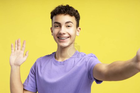 Téléchargez les photos : Portrait de beau garçon souriant, 15 ans influenceur enregistrement vidéo, ayant appel vidéo, portant élégant t-shirt décontracté violet regardant la caméra isolée sur fond jaune. Concept de blogging - en image libre de droit